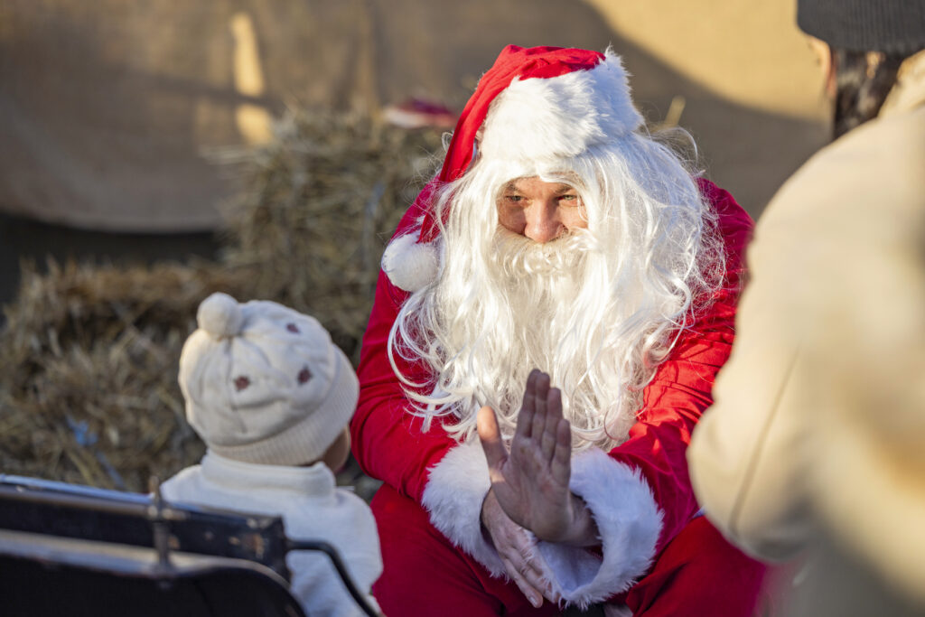 Man utklädd till tomte sitter i släde och gör high-five med ett litet barn, på Stora Sköndals julmarknad.